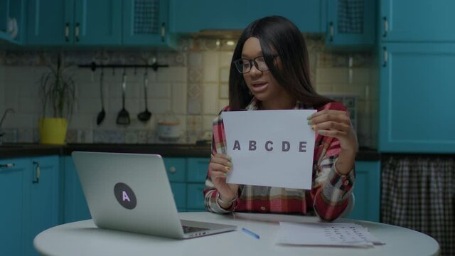 Young African American female online teacher in glasses talking and showing sheet of paper with ABCDE letters looking at laptop. Preschool teacher working remotely from home.