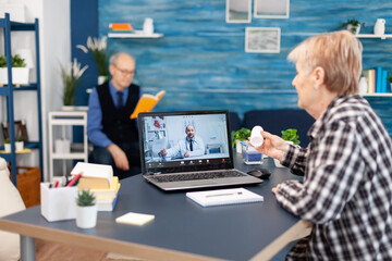 Senior woman showing pills bottle during medical online conference. Elderly woman discussing with healthcare practitioner in the course of remote call and husband is reading a book on sofa.