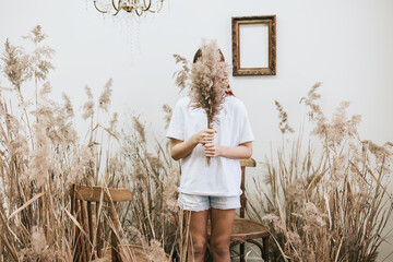 red haired girl closes her face with bouquet wearing a white shirt and denim shorts in a space...