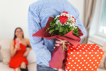 Unexpected moment in routine everyday life Cropped photo of man's hands hiding holding chic bouquet of red roses and gift box behind back, happy woman is on blurred background