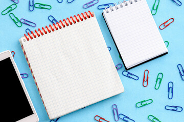 Top view of an office desk with an empty notepad, smartphone, and paper clips on a blue background