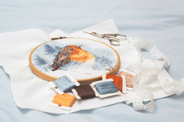 the process of cross stitching a multicolored bird on a white canvas in a wooden hoop, a floss thread, a needle, scissors for needlework
