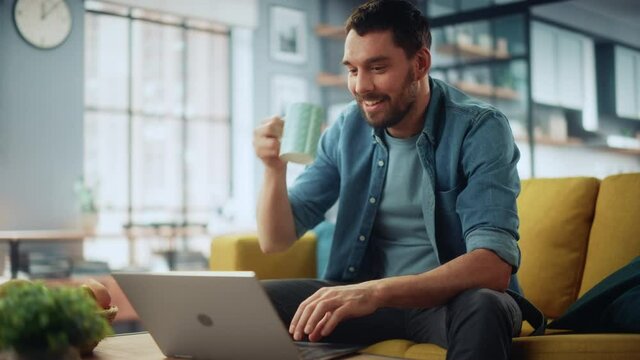 Smiling Caucasian Man Working on Laptop Computer while Sitting on a Sofa Couch in Stylish Cozy Living Room. Happy guy Having Fun at Home. Browsing Internet, Drinking Coffee from a Mug.
