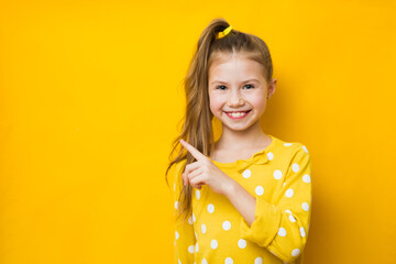Young beautiful girl points index finger aside and demonstrates copy space for your advertisement on yellow background. Look your best promo here