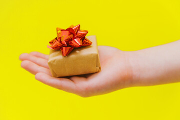 A child's hand holds a small gift box with a red bow on a yellow background. A birthday present.