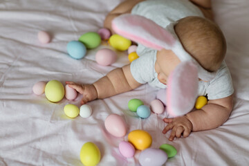 happy caucasian baby girl six months old wearing bunny ears headband, lying on bed at home in bedroom with colored easter eggs
