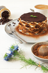 Round tiramisu, homemade no-bake traditional Italian sweet dessert. Blue table with cake, bowl with cocoa powder, round sponge biscuits, coffee beans and coffee with milk. White background.