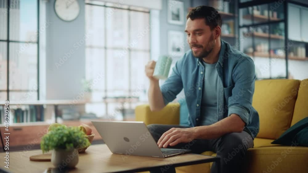 Poster smiling caucasian man working on laptop computer while sitting on a sofa couch in stylish cozy livin