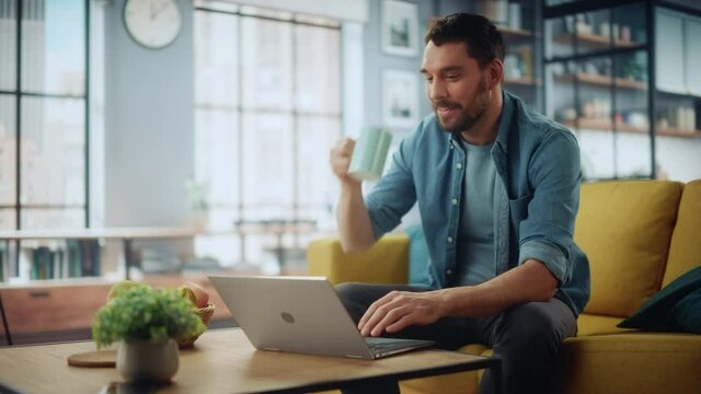 Smiling Caucasian Man Working on Laptop Computer while Sitting on a Sofa Couch in Stylish Cozy Living Room. Happy guy Having Fun at Home. Browsing Internet, Drinking Coffee from a Mug.