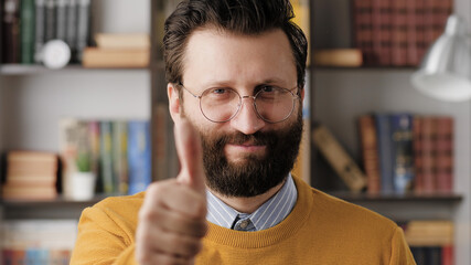 Obraz premium Man thumb up. Positive smiling bearded man in glasses in office or apartment room looking at camera and shows his thumb up. Close-up