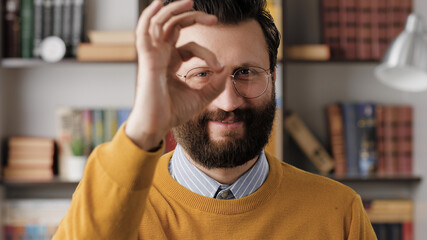 Obraz premium Man shows OK. Positive smiling bearded man in glasses in office or apartment room looks at camera and shows OK gesture with his fingers. Close-up
