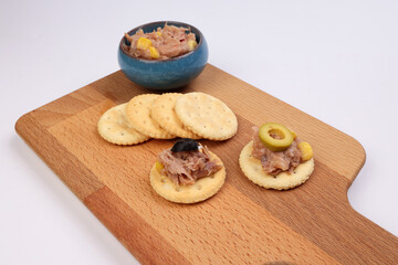 Tuna fish spread topping cracker biscuit tapas corn green black olive small bowl on wooden board over white background