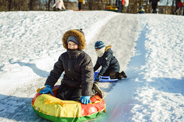 two cute caucasian boys sledding down the icy slope in park on snow saucer and tubing. Winter activities concept. Happy childhood. Image with selective focus.