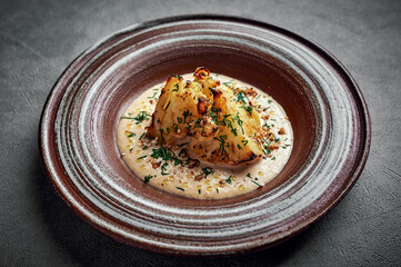Baked homemade cauliflower in gruyere cheese sauce, herbs and olive oil in a ceramic plate on a dark background, healthy food, copy space, top view