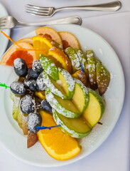 Sliced fruit (orange, pear, Apple, grapes ) sprinkled with powdered sugar, plastic skewers on a white plate close-up and metal forks on a white tablecloth background