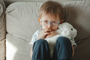 A child with autism in glasses sits on the sofa and sad, hug a s