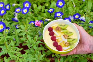 Rolgordijnen Mango banana pineapple smoothie bowl topped with raspberry, kiwi, walnut and coconut chips. Flower background © AmalliaEka