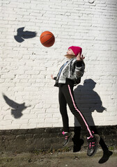 A sporty girl plays with a basketball against the background of a white brick wall with the shadow of a bird.