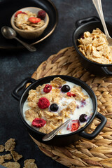 Corn flakes with strawberry and blueberry in black bowls over dark background, top view.