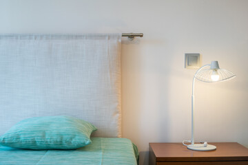 Close up of top of bed with a pillow and a nightstand with a lit lamp on top. Minimalist design.