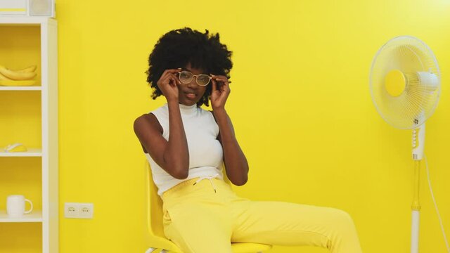Creative portrait of a confident African woman, model, sitting on yellow chair next to a fan, chilling out and smiling, putting on glasses, yellow interior, Foreground, Slow motion.