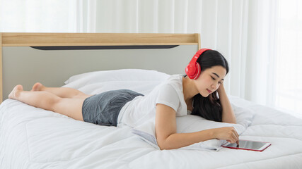 Youn and beautiful asian woman wearing red headphone laying on bed in the bedroom and using tablet computer to work and connect to internet
