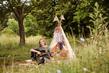 Authentic woman is playing the guitar next to wigwam outdoors in summer. © Natalia