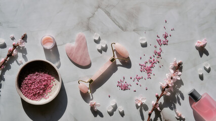 Crystal rose quartz facial roller and Gua sha stone for beauty facial massage therapy, Flat lay on marble table with magnolia flowers. Long shadows and candle. Essential oil bottle. Panoramic image. - obrazy, fototapety, plakaty