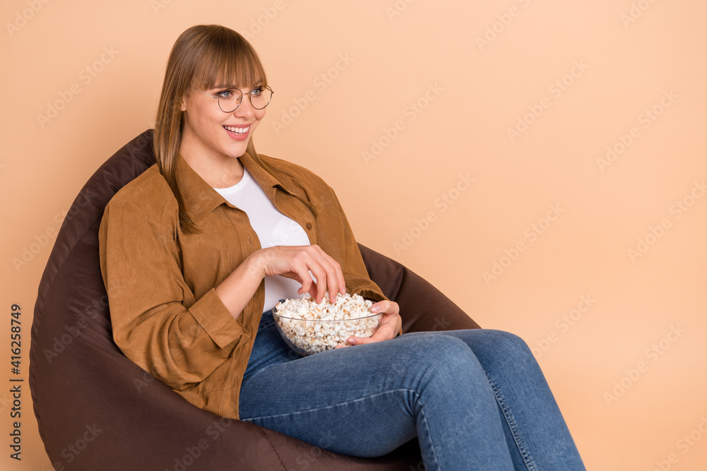 Poster Photo of positive pretty young woman look empty space tv eat popcorn isolated on pastel beige color background