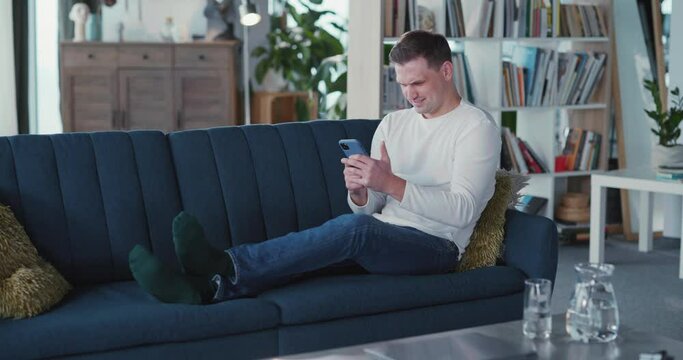 Young manusing smartphone in living room casual modern apartment texting messages netowkring on social media pps indoors.