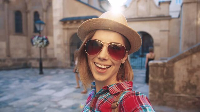 Close up of young beautiful woman tourist in sunglasses and hat posing at camera standing in city center. View from camera.