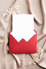 Red envelope with blank paper card inside on beige satin. Flat lay, top view, copy space. Wedding invitation card mockup.