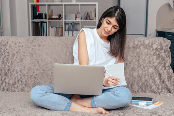 A dark-haired mixed race girl is sitting at home on the couch,using a laptop and making notes in a notebook.Freelancer working, online education,distance working,business online concept.