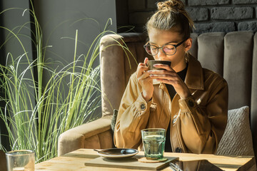 Young woman drinking coffee cup and sitting in cafe. Girl in glasses pauses after work.
