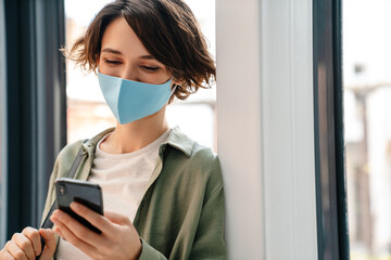 Happy girl in protective mask using cellphone while leaning on wall