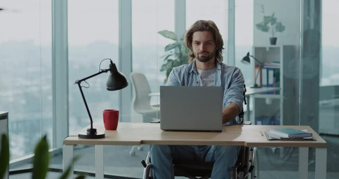 Young caucasian handsome disabled man in wheelchair sitting by desk using computer laptop for work. Business center. Corporate person.