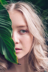 Naklejka premium Portrait of a blonde teenage girl, covering half of her face with a plant leaf.Concept of closeness to nature.Eco,urban jungle,biophilia concept.Selective focus.