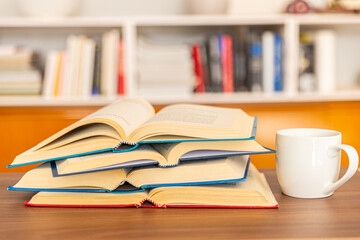 Open books, arranged one on top of the other, with a white cup of tea or coffee next to them, on a wooden surface, in front of the bookcase. Reading, studying, learning.