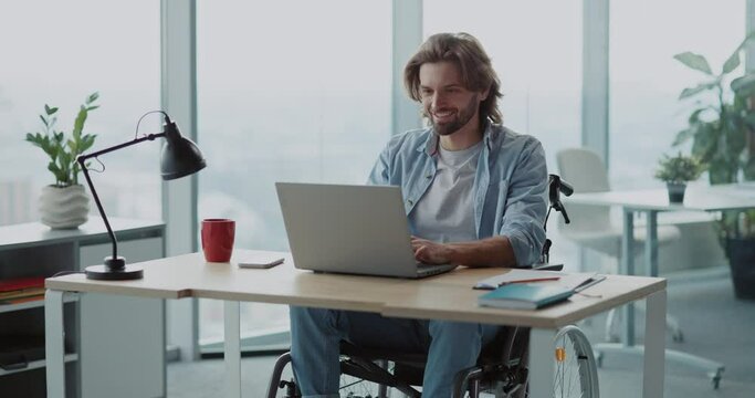 Young handsome disabled man in wheelchair using computer laptop device technology in corporate company office.