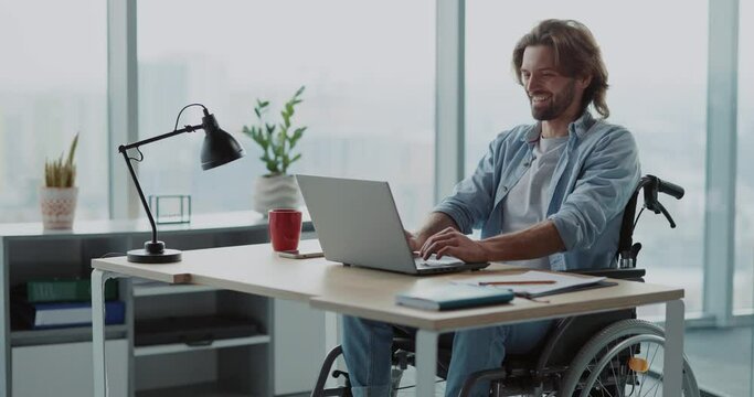 Handsome young disabled man in black wheelchair sitting by computer desk using portable laptop reading news doing work alone in the office.