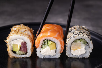 Sushi rolls set with avocado, cheese, salmon and tuna on black background. Chopsticks taking the sushi roll.