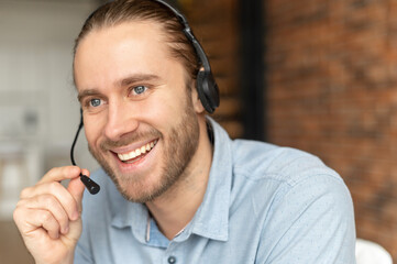 Handsome European man with headset looking away and talking, has a light brown hair, gray eyes and beard, smiling friendly, operator call-center, confident, wearing blue shirt, at the modern office