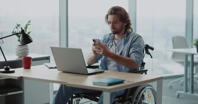 Handsome young man in wheelchair sitting on break using mobile phone typing messages communicating online wirelessly in corporate office.