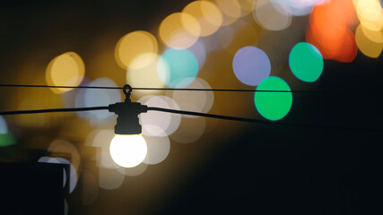 A bright light bulb shines with a warm light with a black wire against the backdrop of a beautiful, bustling city at night. Car traffic in shallow depth of field. Street light. Twinkling lights, bokeh