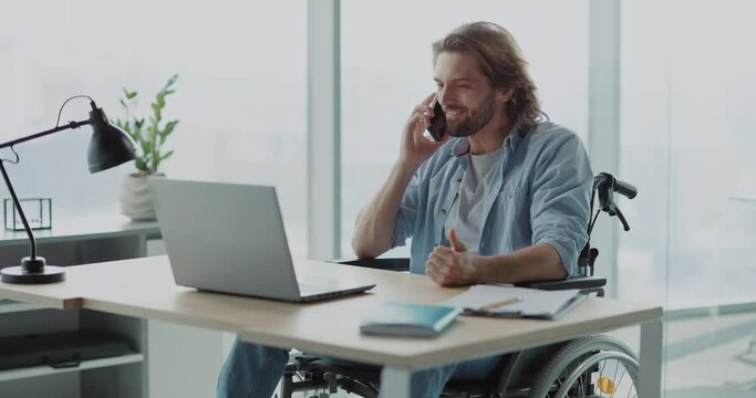 Optimistic caucasian young man with long hair sitting in corporate office talking on mobile phone. Dialing cellphone. People with disabilities.