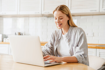 Joyful blonde girl smiling and using laptop while sitting at kitchen