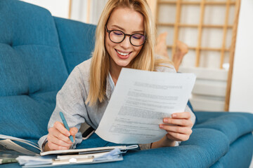 Happy student girl in eyeglasses doing homework while lying on couch