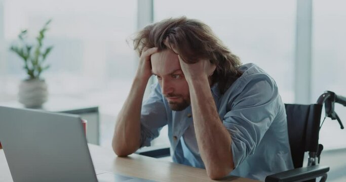 Young depressed office employee disabled in wheelchair sitting on corporate office workspace worrying about working duties. Frutstration. Emotions.