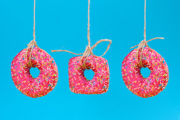 Three doughnuts of different shapes with pink frosting, suspended on a string. Close-up. Blue background