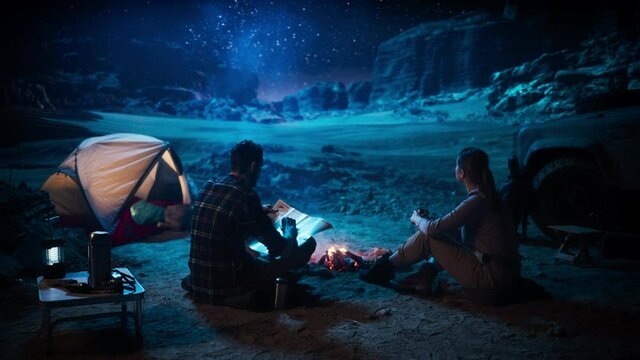 Happy Couple of Hikers Camping in the Canyon at Night Together, Sitting by Campfire, Plan Hiking Trekking Journey Route with a Map. Two Traveling people Talk, Milky Way Star Watching. Vacation Trip